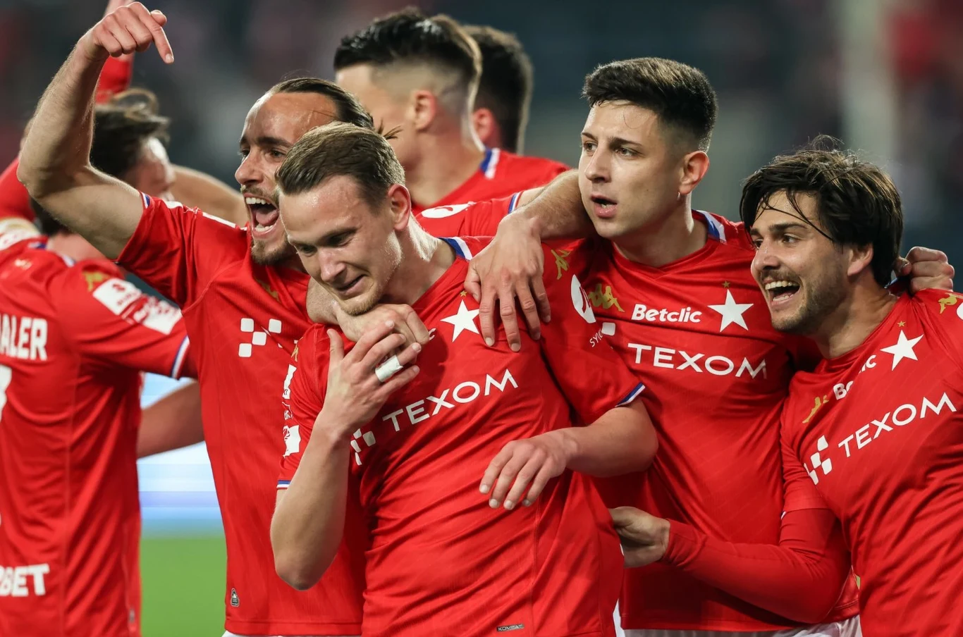 Wisła Kraków Grupa piłkarzy w czerwonych koszulkach z logo sponsorów świętuje razem zdobycie bramki, obejmując się i wyrażając radość.