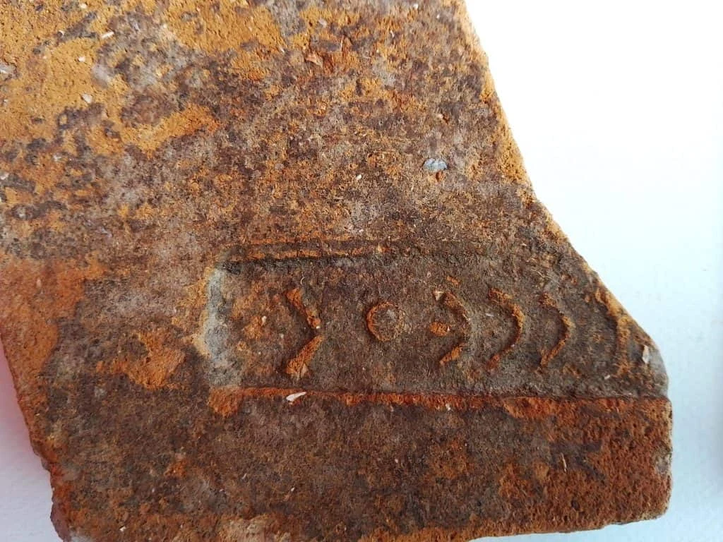 Fragment starego, zardzewiałego metalu z wyrytymi geometrycznymi znakami przypominającymi groty strzał i kropkę w środku prostokątnej ramki.