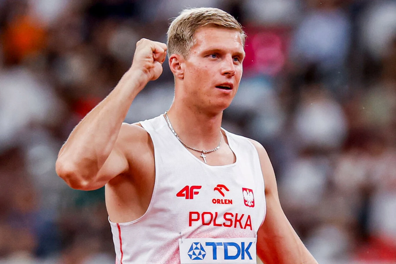 Lekkoatleta w biało-czerwonym stroju z napisem 'Polska' z zaciśniętą pięścią w geście zwycięstwa na tle stadionu lekkoatletycznego.