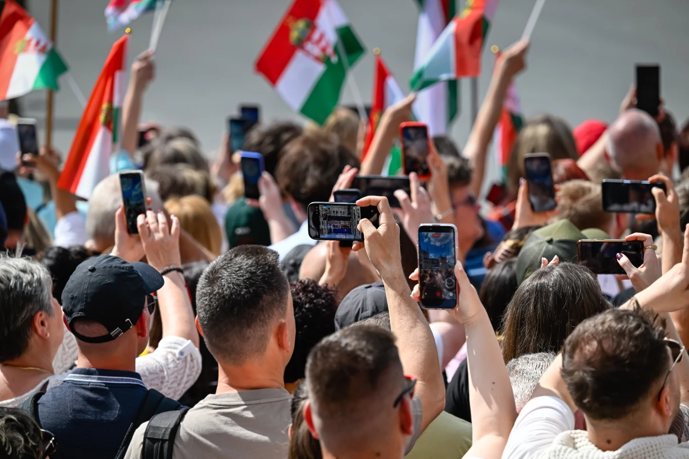Tłum ludzi trzymających telefony komórkowe skierowane do przodu oraz ręce powiewające flagami w dominujących barwach czerwieni, bieli i zieleni.