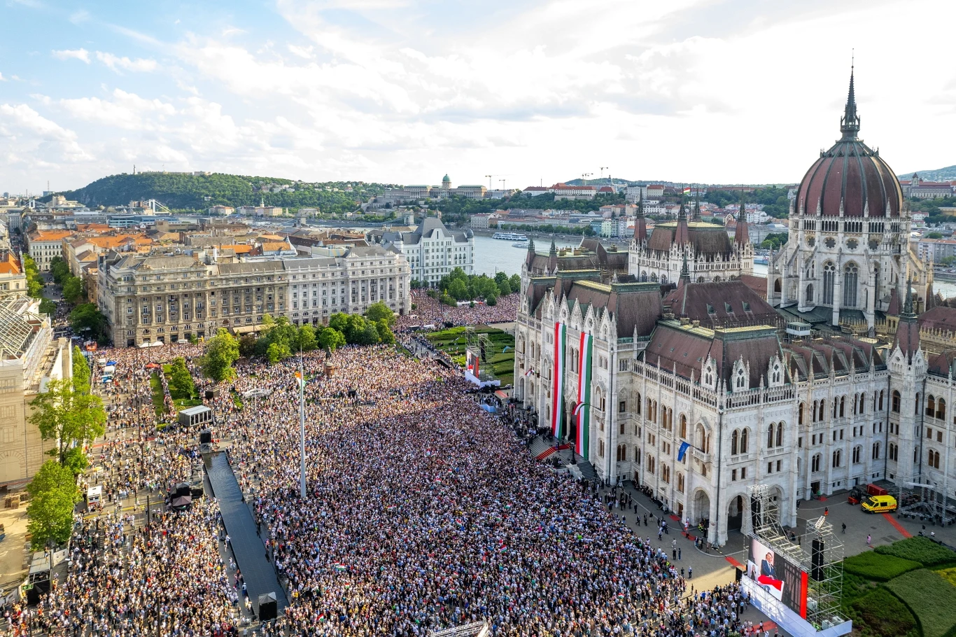 Ogromny tłum ludzi zgromadzony przed budynkiem parlamentu w Budapeszcie, obok którego powiewają duże flagi w narodowych barwach Węgier; w tle panorama miasta oraz Dunaj.
