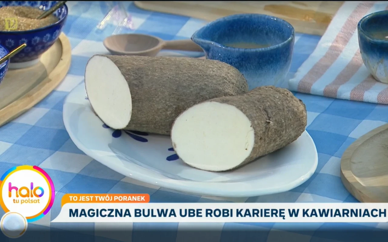 Przekrojona na pół bulwa ube o fioletowej skórce i białym wnętrzu leży na owalnym, białym talerzu na niebiesko-białym obrusie, w otoczeniu niebieskiej i beżowej zastawy stołowej.