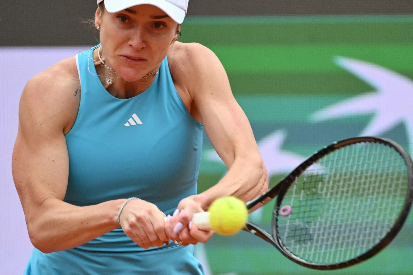 Kobieta w niebieskiej sportowej koszulce i białej czapce odbija piłkę tenisową rakietą, koncentrując się na wykonaniu uderzenia.