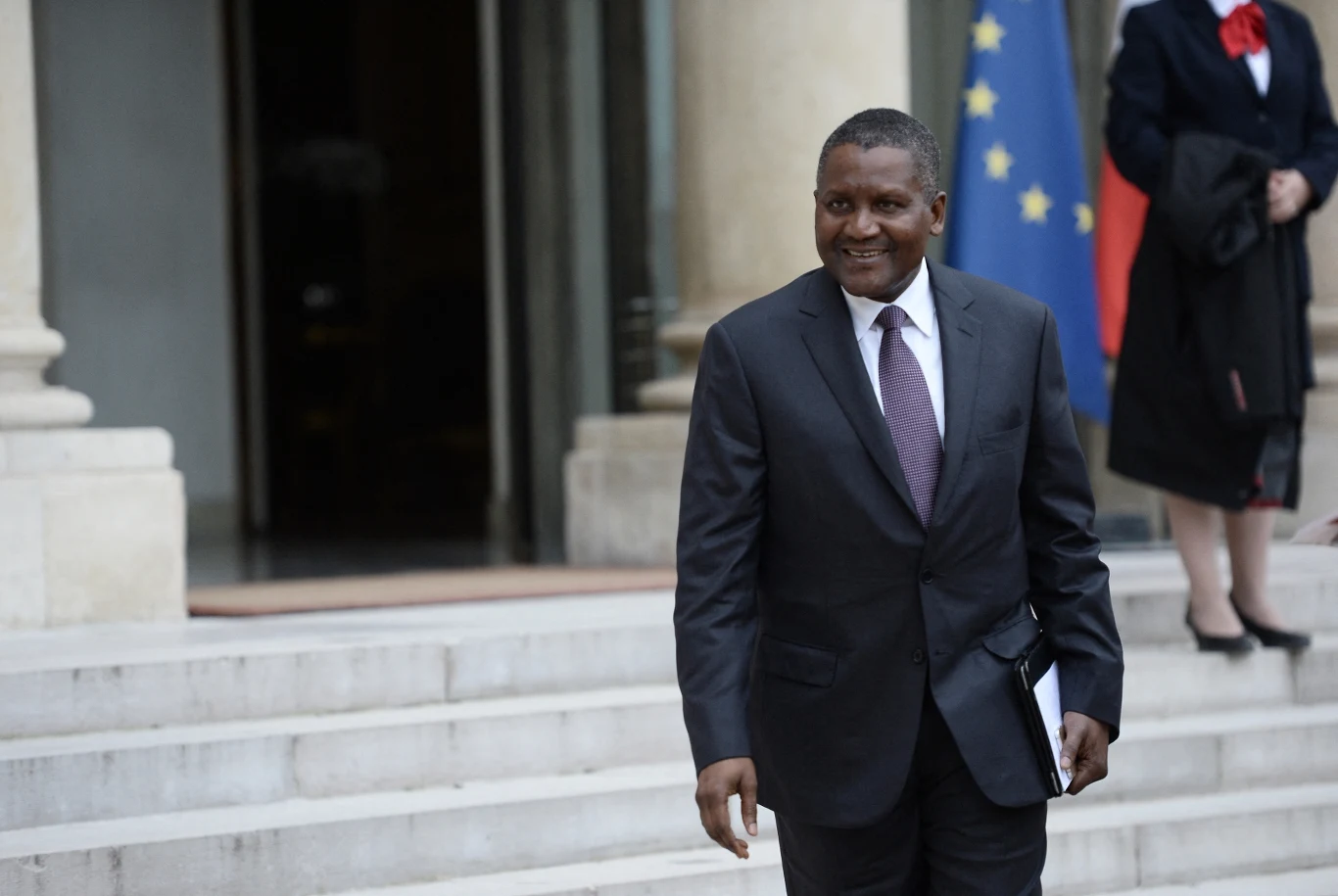 Najbogatszy człowiek Afryki rozważa budowę nowej rafinerii ropy naftowej w Kenii Aliko Dangote w garniturze stoi na schodach przed budynkiem z flagą UE w tle, trzymając dokumenty