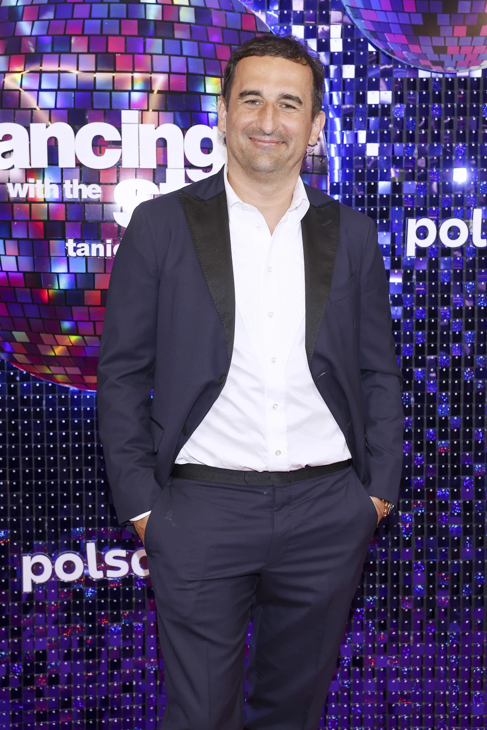 Michał Koterski Mężczyzna w granatowym garniturze i białej koszuli pozuje na tle błyszczącej ścianki ozdobionej kolorowymi mozaikami oraz logo programu Dancing with the Stars.