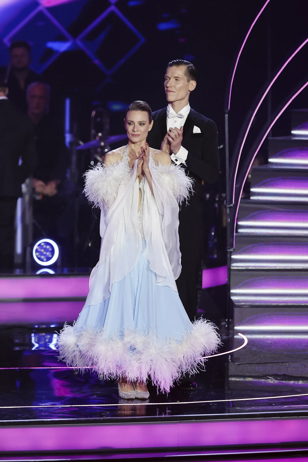 Magda Boczarska i Jacek Jeschke Tancerka w eleganckiej, błękitnej sukni ozdobionej piórami stoi przodem, a za nią partner w czarnym fraku i białej koszuli, oboje klaszczą znajdując się na oświetlonej scenie tanecznej.