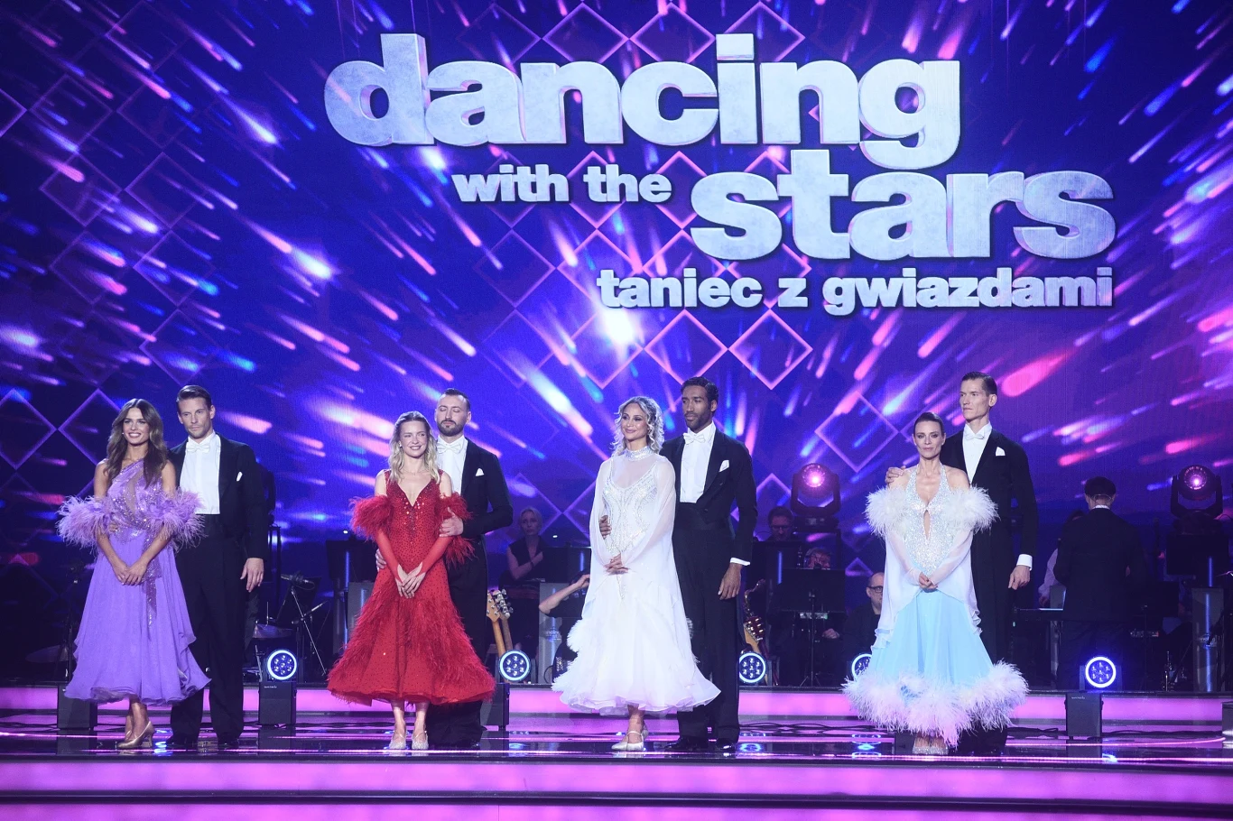 Finał Tańca z gwiazdami Pary taneczne w eleganckich kreacjach stoją na kolorowo oświetlonej scenie programu 'Dancing with the Stars. Taniec z gwiazdami' na tle dużego napisu z nazwą show.