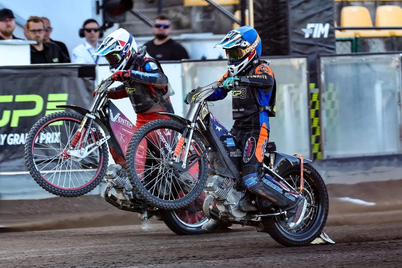 Dwóch żużlowców w kolorowych kombinezonach i kaskach ściga się na motocyklach z uniesionymi przednimi kołami, wzbudzając kurz na torze stadionu.