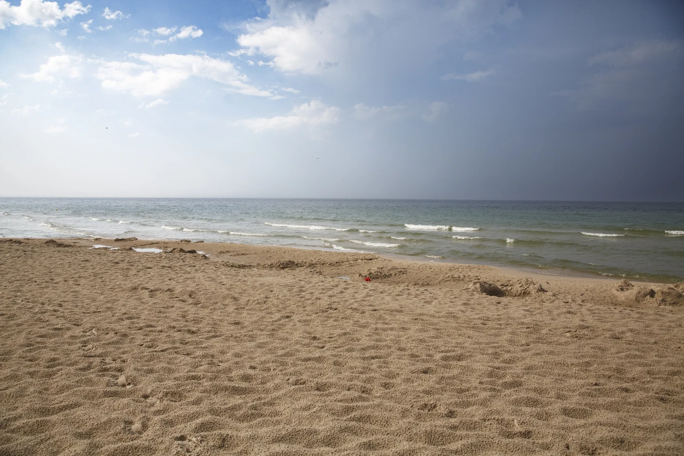Piaszczysta plaża rozciągająca się wzdłuż spokojnego morza, nad którym unosi się częściowo zachmurzone niebo.