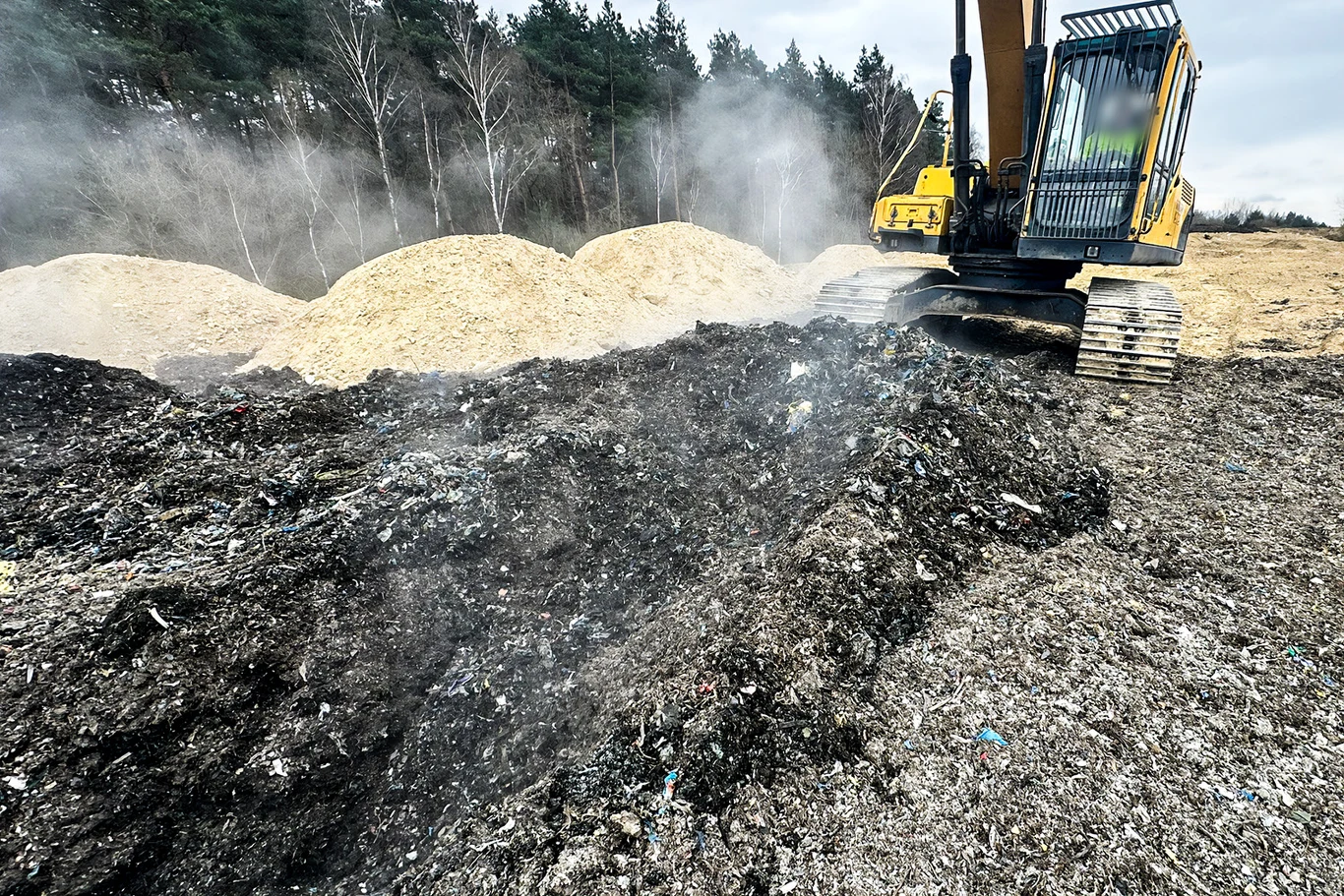 Koparka pracująca na dymiącej hałdzie odpadów, w tle żółtawe kopce trocin i pas lasu.