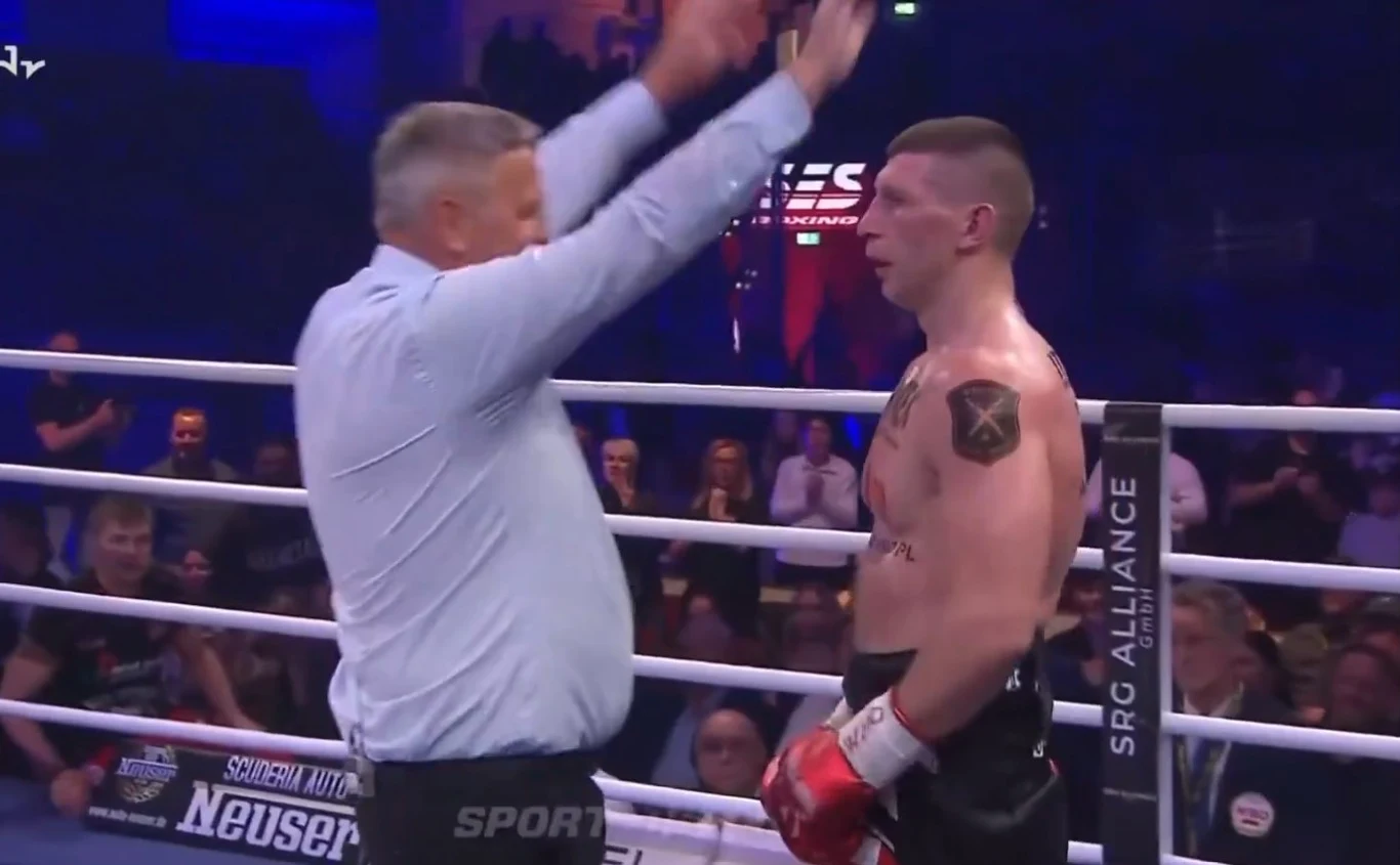 Sędzia podnosi ręce przed bokserem stojącym w ringu, wokół widownia i światła areny.