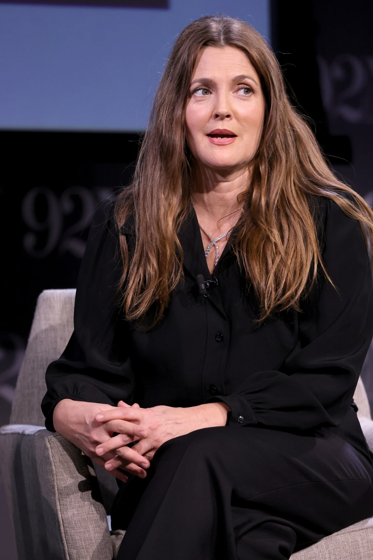 Drew Barrymore pokazuje regularnie swoją twarz bez makijażu w mediach społecznościowych Kobieta o długich, rozpuszczonych włosach w ciemnym stroju, siedząca na jasnym fotelu z dłońmi złożonymi na kolanach, patrząca w bok podczas rozmowy na scenie.