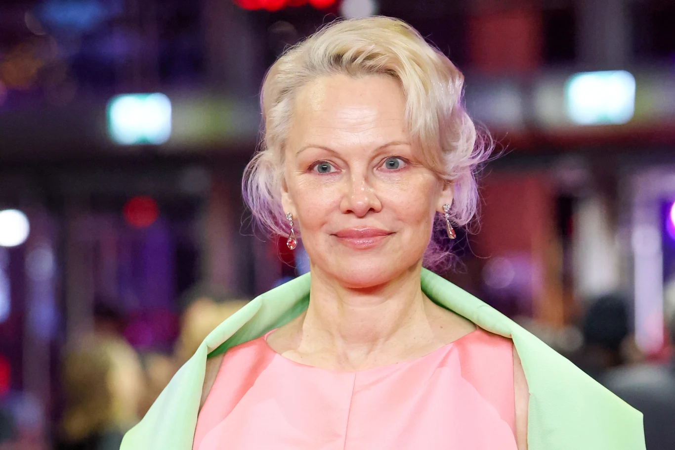 Pamela Anderson od dłuższego czasu promuje bardzo naturalny look Kobieta o jasnych włosach spiętych do góry, ubrana w różową sukienkę i zieloną pelerynę, stoi na tle rozmytego wnętrza z kolorowymi światłami.