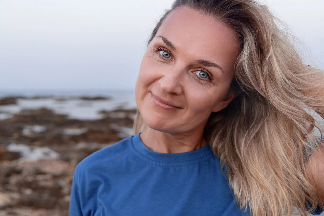 Najbardziej spontaniczne znaki zodiaku. Decyzje podejmują w kilka sekund Uśmiechnięta kobieta z blond włosami ubrana w niebieską koszulkę stoi na tle skalistego wybrzeża.