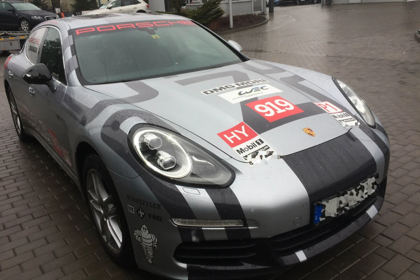 Srebrny samochód sportowy Porsche z wyraźnymi naklejkami rajdowymi, w tym numerem 919 i oznaczeniami partnerów, zaparkowany na brukowanej nawierzchni.