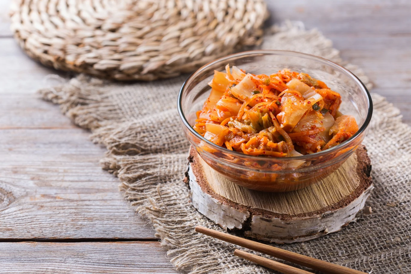 Miseczka kimchi z wyraźnie widocznymi kawałkami piklowanej kapusty i przyprawami, ustawiona na krążku drewna, obok leżą drewniane pałeczki.