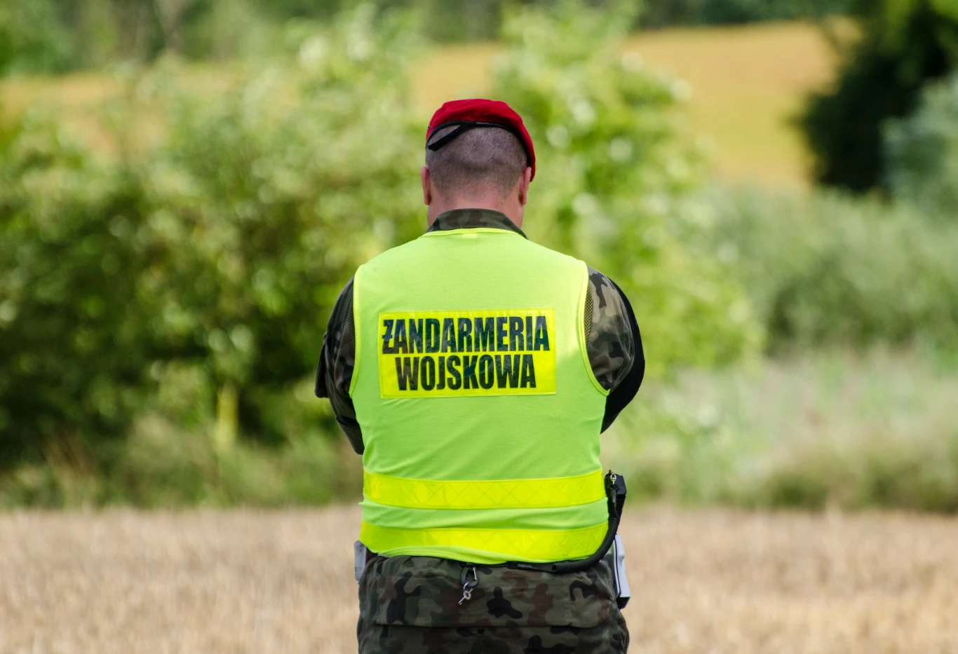 Przy granicy z Rosją w sobotę znaleziono drona (zdj. ilustracyjne) Żołnierz Żandarmerii Wojskowej w żółtej kamizelce stoi tyłem na polu.