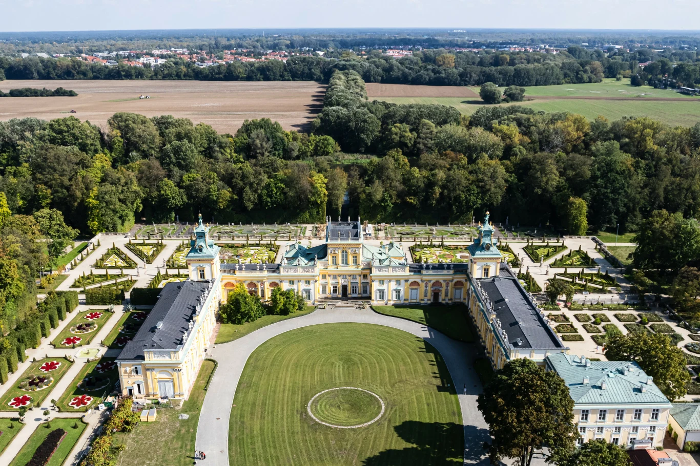 Barokowy pałac otoczony symetrycznie zaprojektowanymi ogrodami, z centralnym dziedzińcem i szerokimi aleiami, w tle rozległe połacie zieleni i pola uprawne.