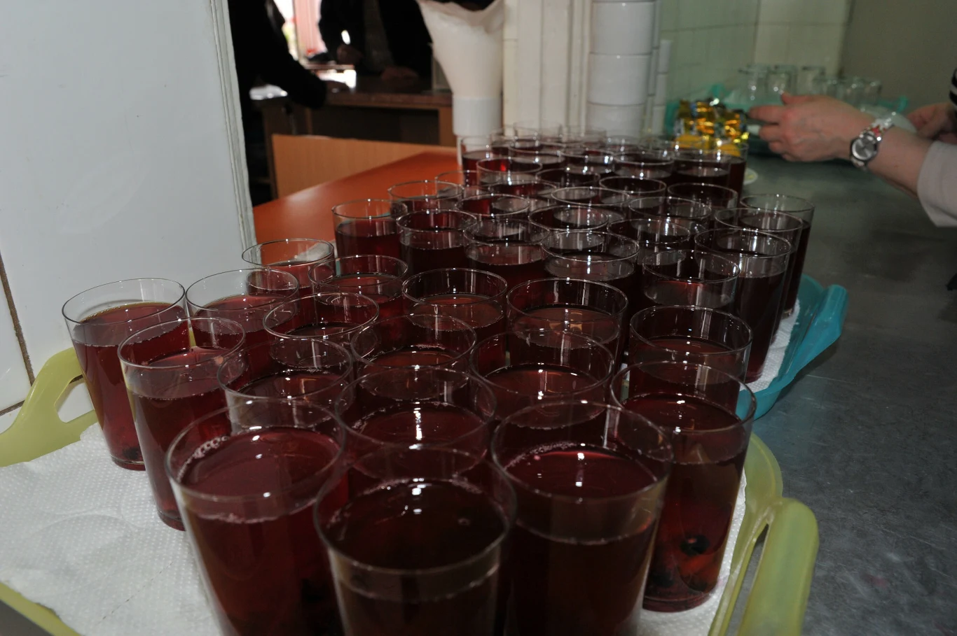 Rzędy plastikowych kubków wypełnionych czerwonym napojem ustawione na tacy na metalowym blacie.