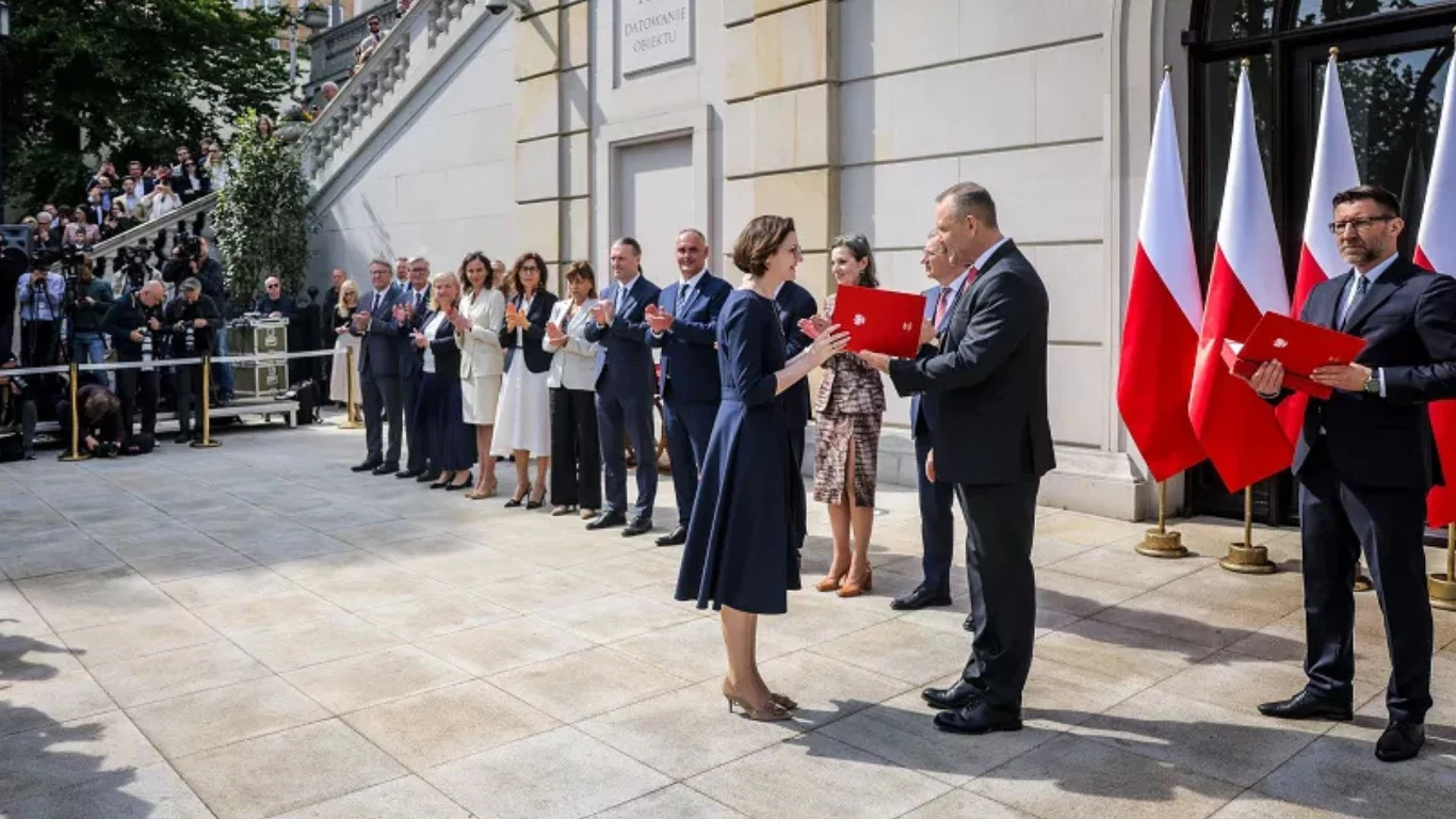 Uroczystość wręczenia dokumentu między kobietą a mężczyzną na tle trzech ustawionych obok siebie flag Polski, w otoczeniu elegancko ubranych osób stojących w szeregu i publiczności na schodach.