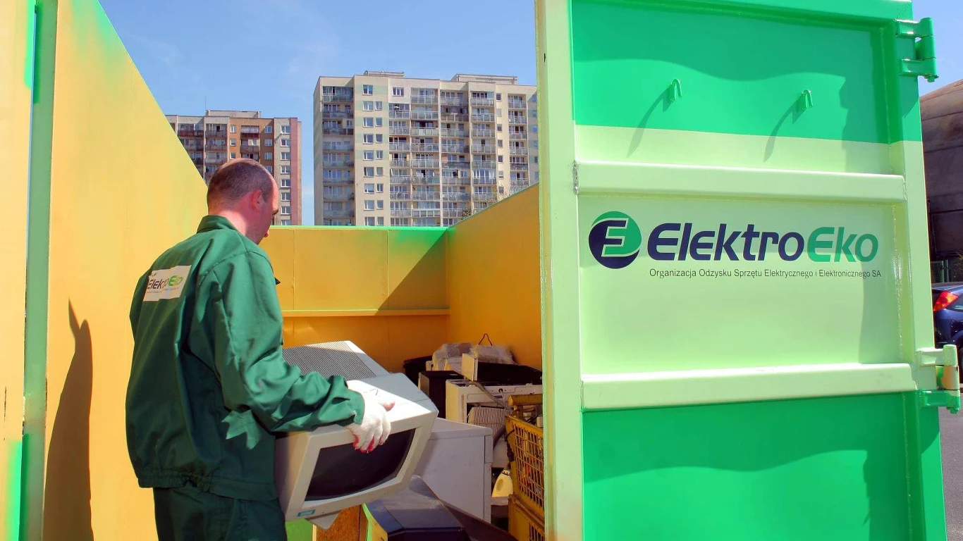 Mężczyzna w zielonym uniformie wrzuca stary monitor komputerowy do kontenera oznaczonego logo ElektroEko, w tle widoczne są bloki mieszkalne.