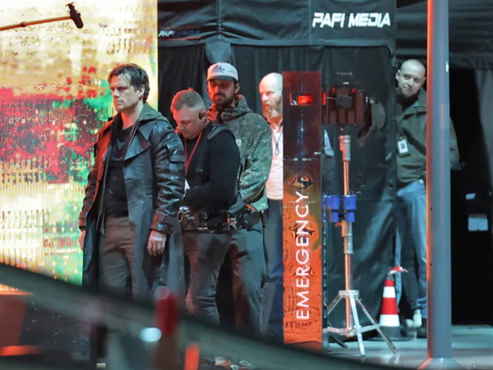 Mężczyzna w ciemnym płaszczu stoi obok kilku osób z ekipy technicznej na tle oświetlonej ściany i oznaczenia „EMERGENCY”.