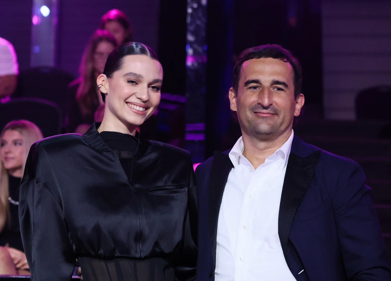 Elegancko ubrana kobieta w czarnej sukni stoi obok mężczyzny w granatowej marynarce i białej koszuli, oboje uśmiechnięci, w otoczeniu siedzących w tle osób.