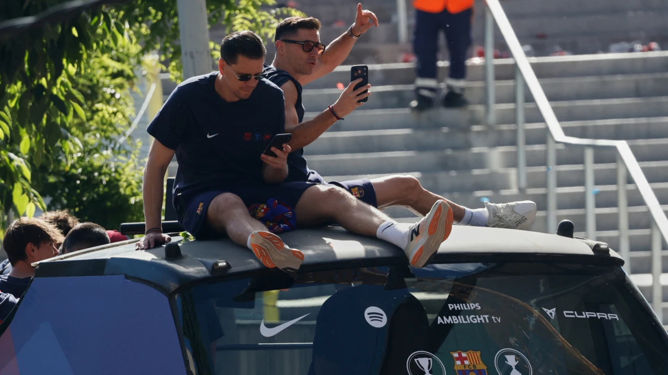 Dwóch mężczyzn w sportowych ubraniach siedzi na dachu pojazdu, robiąc sobie zdjęcia smartfonami, jeden z nich pokazuje gest uniesionego kciuka.