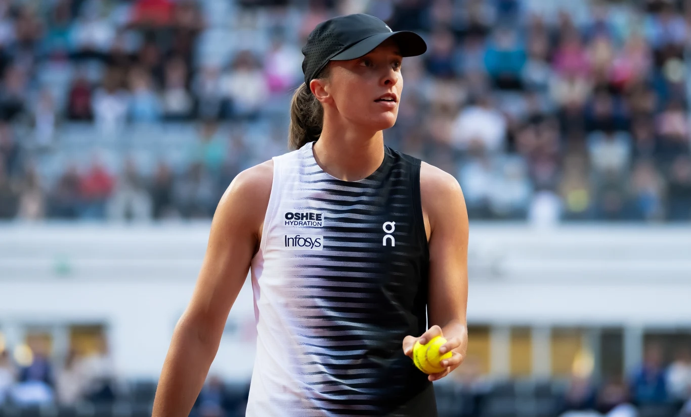 Tenisistka w sportowej koszulce i czapce trzymająca piłkę tenisową podczas meczu na korcie, w tle widownia.