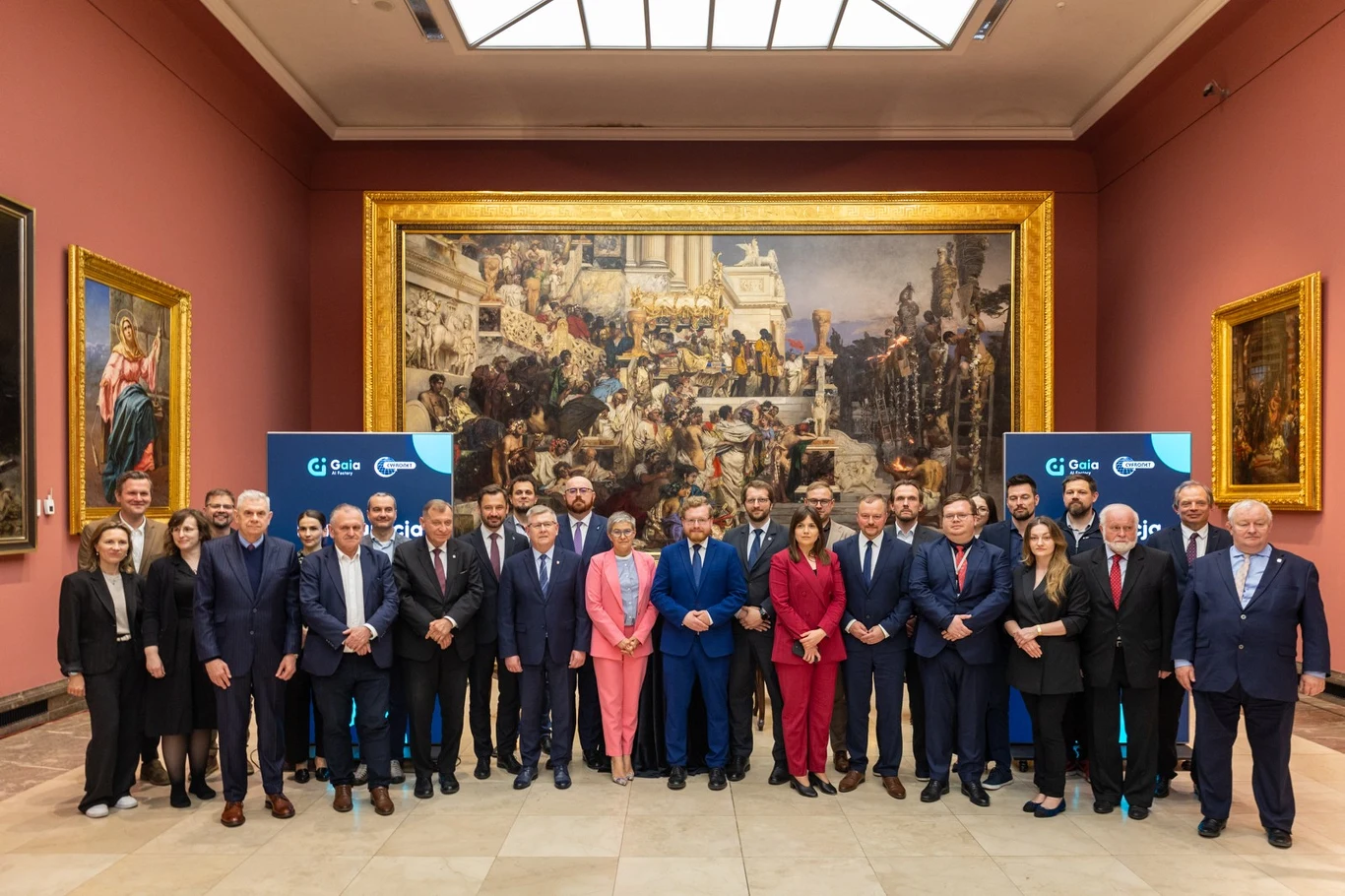 Grupa formalnie ubranych osób stojąca na tle wielkiego obrazu historycznego w muzealnym wnętrzu, z dwoma niebieskimi banerami po bokach.