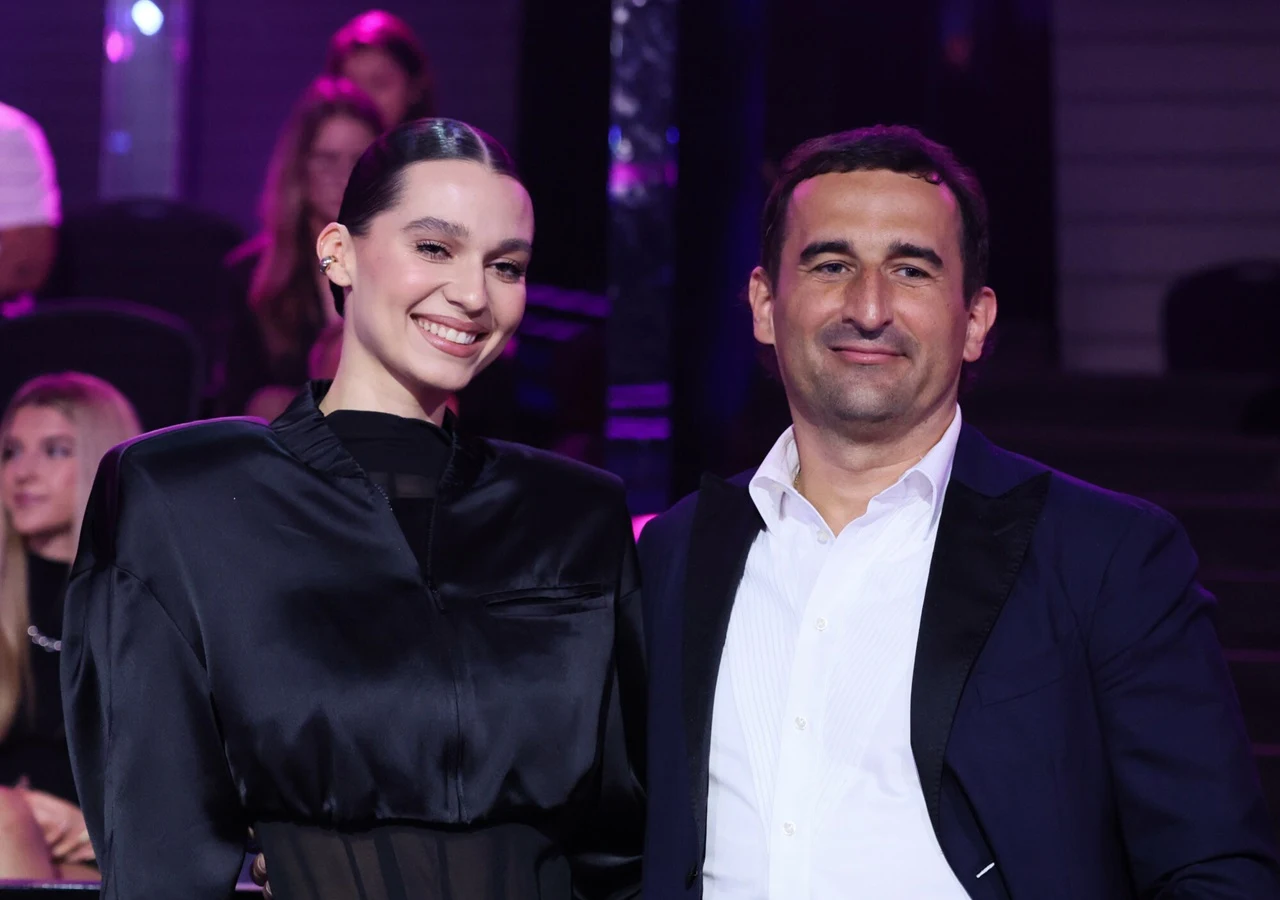Uśmiechnięta kobieta w eleganckiej czarnej bluzce i mężczyzna w granatowej marynarce oraz białej koszuli stoją obok siebie, otoczeni przez siedzących w tle ludzi na widowni oświetlonej fioletowym światłem.