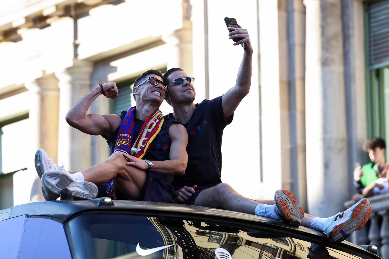 Dwóch mężczyzn w sportowych strojach siedzi na dachu pojazdu, pozując do wspólnego selfie; jeden z nich owinięty w szalik w barwach klubowych uśmiecha się i prezentuje napięty biceps.