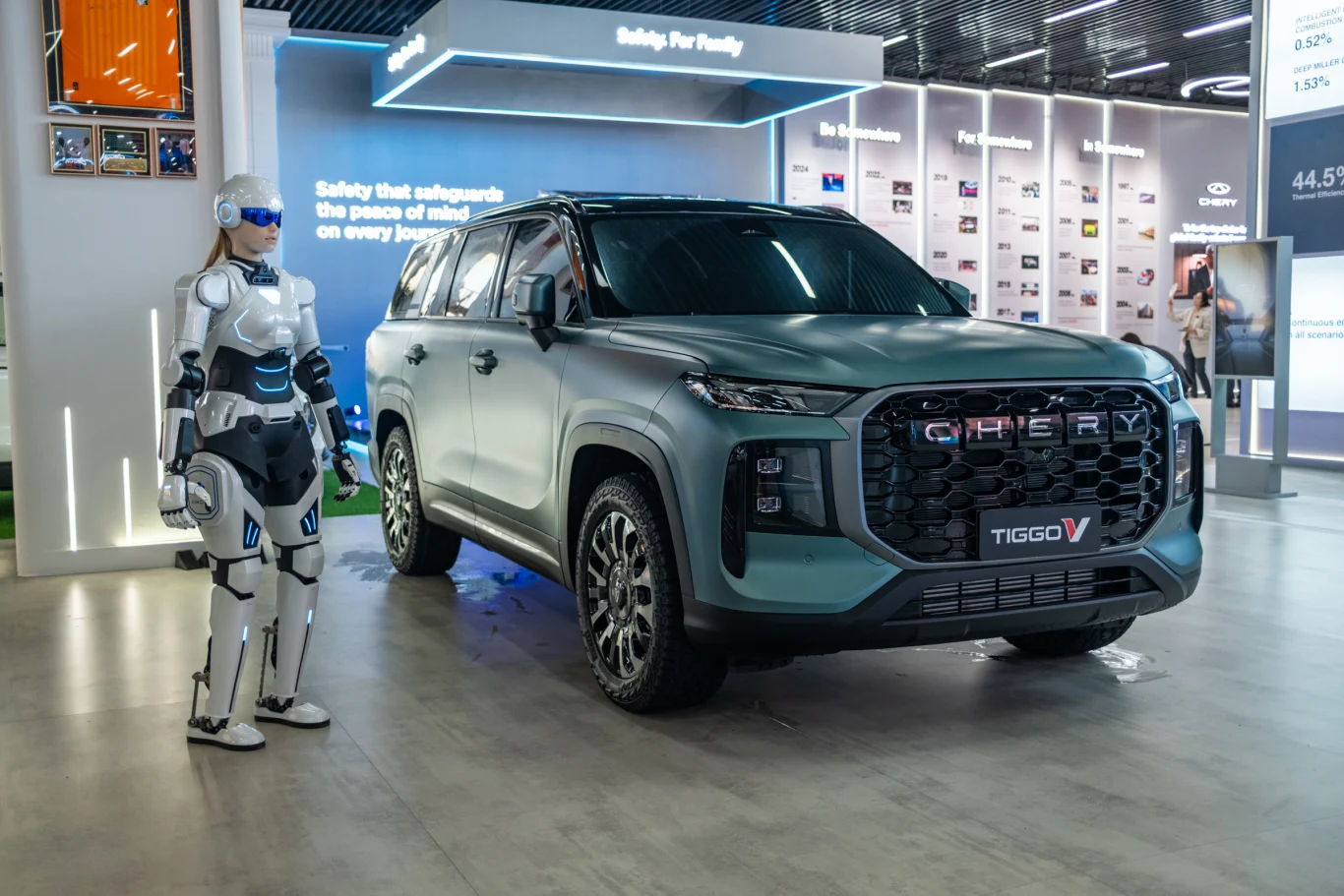 Nowoczesny SUV Chery Tiggo 8 Pro V ustawiony w jasnej hali targowej, obok którego stoi humanoidalny robot w futurystycznej zbroi.