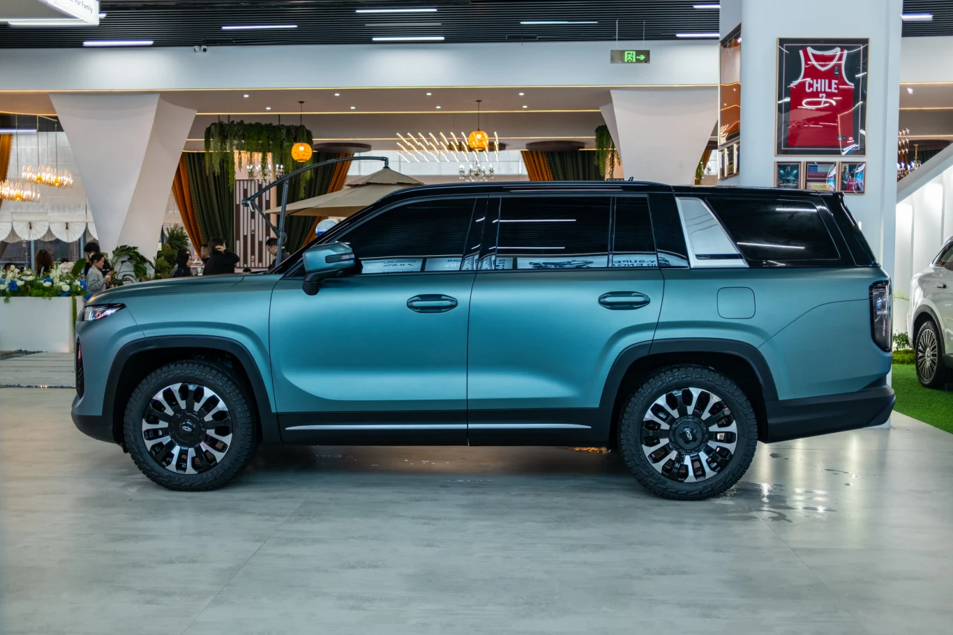 Duży, nowoczesny SUV w matowym, niebiesko-zielonym kolorze z czarnymi akcentami, masywnymi kołami i przyciemnianymi szybami, zaparkowany na eleganckiej hali wystawowej.