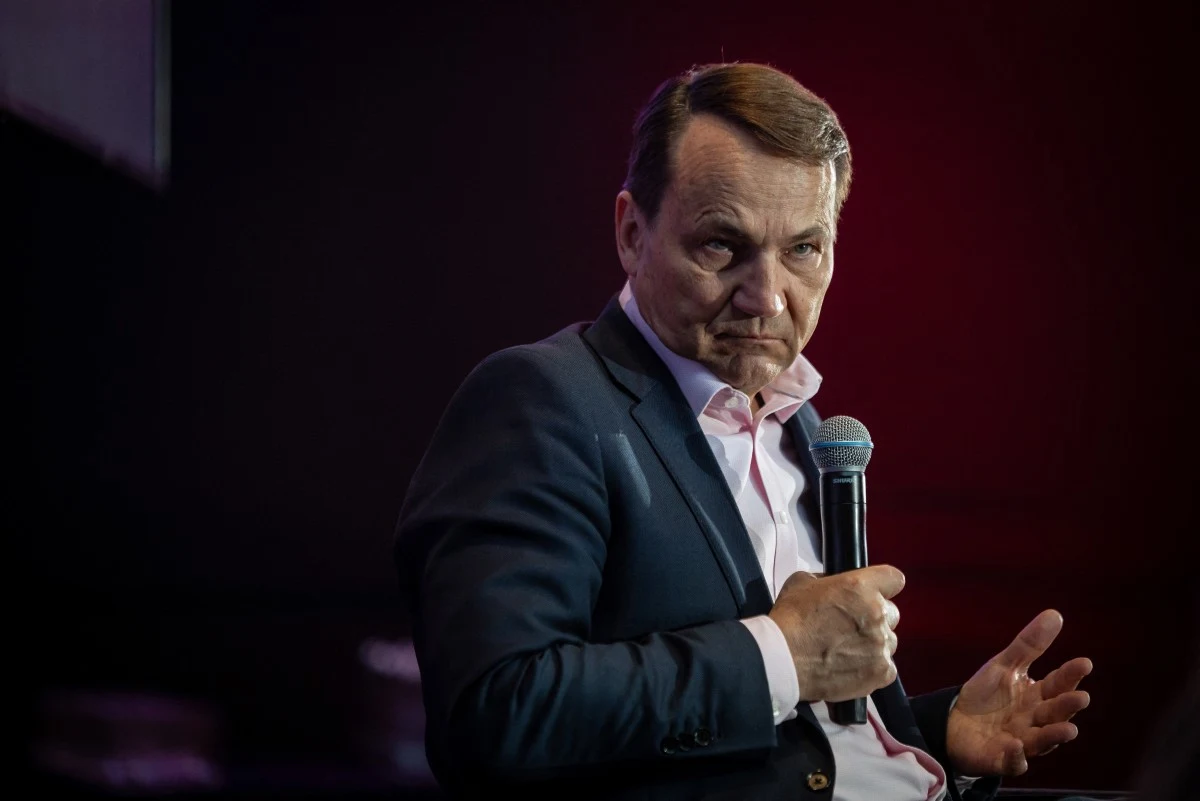 Mężczyzna w garniturze z mikrofonem w dłoni siedzi na scenie, wpatrzony poważnie w kierunku publiczności.
