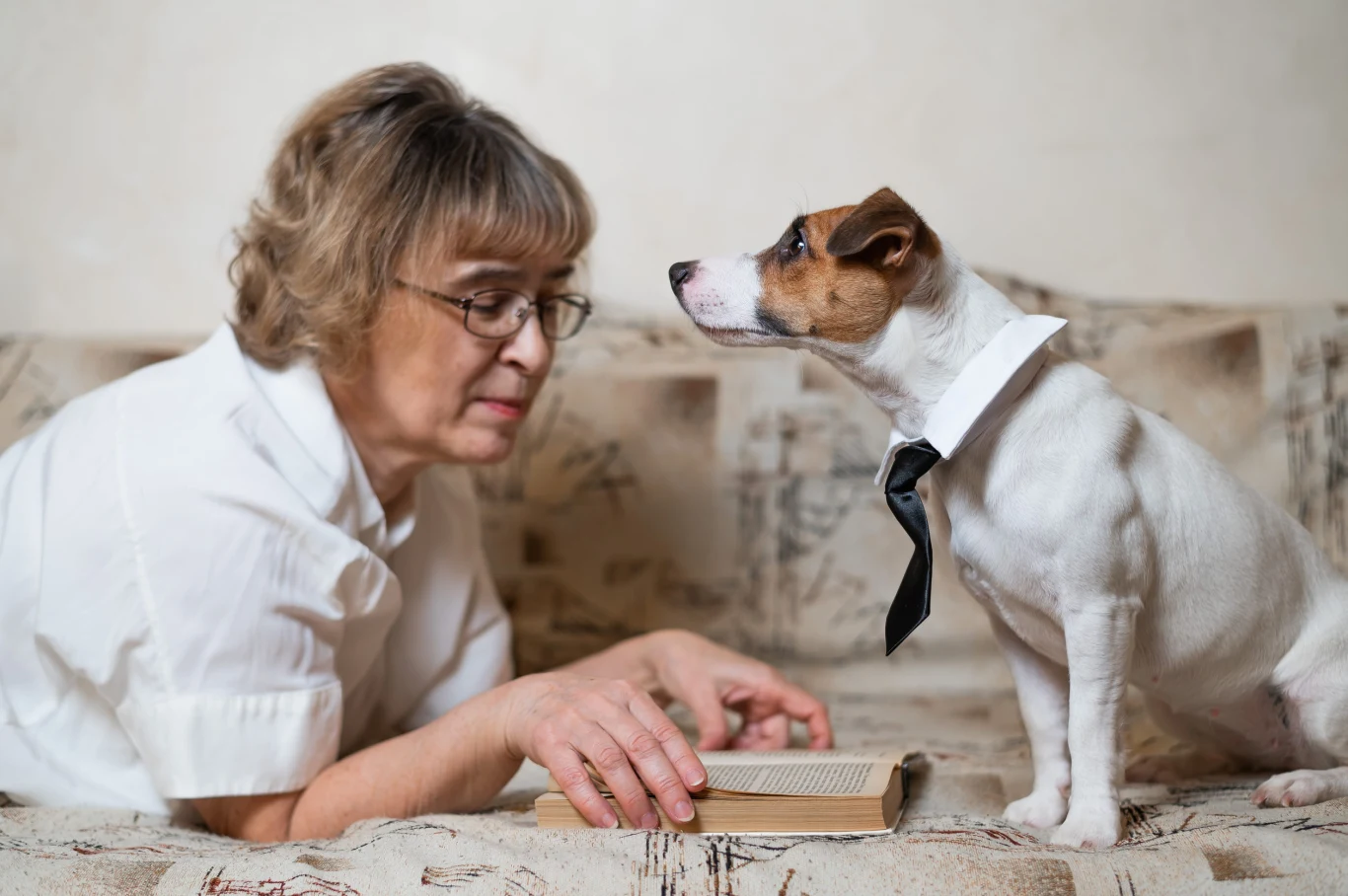 Samotność seniorów to coraz częstszy problem. Zwierzę może wiele zmienić Starsza kobieta w okularach leży na kanapie z książką, obok niej pies w białej koszuli i czarnej krawacie.