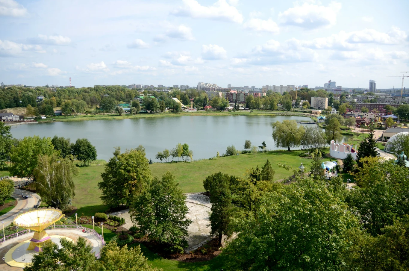 Duże jezioro otoczone zielenią, z licznymi drzewami oraz parkiem w pierwszym planie, za którym rozciąga się panorama miasta.