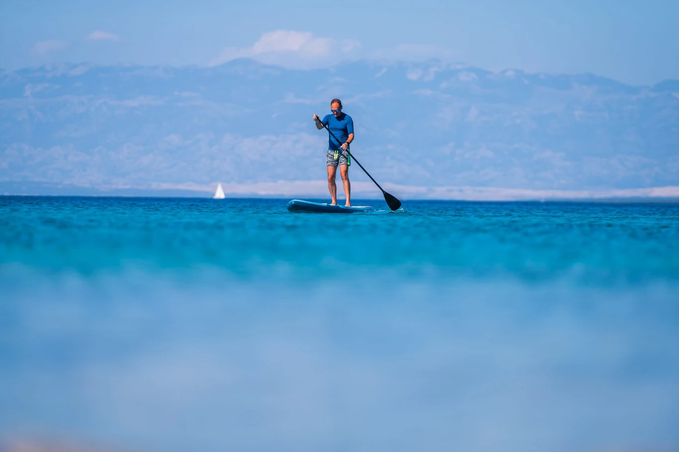Mężczyzna w niebieskiej koszulce i krótkich spodenkach stoi na desce do paddleboardingu, trzymając wiosło na spokojnej, błękitnej wodzie z górami w tle.