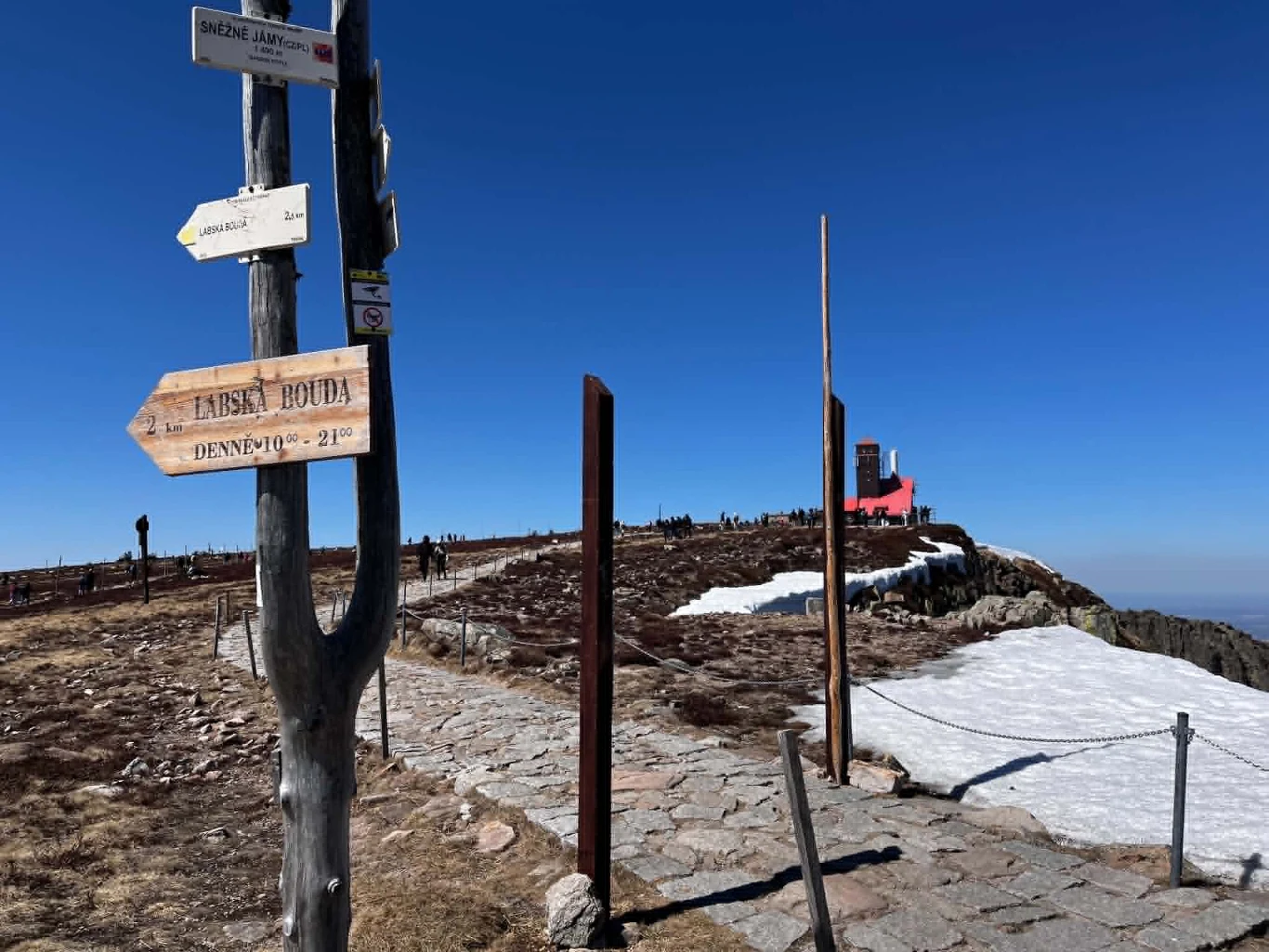 Szlak wiedzie wzdłuż granicy polsko-czeskiej Drewniany słupek z kierunkowskazami przy kamiennej ścieżce prowadzącej przez górski szczyt z resztkami śniegu, w oddali charakterystyczny czerwony budynek oraz pojedyncze sylwetki turystów na tle czystego nieba.