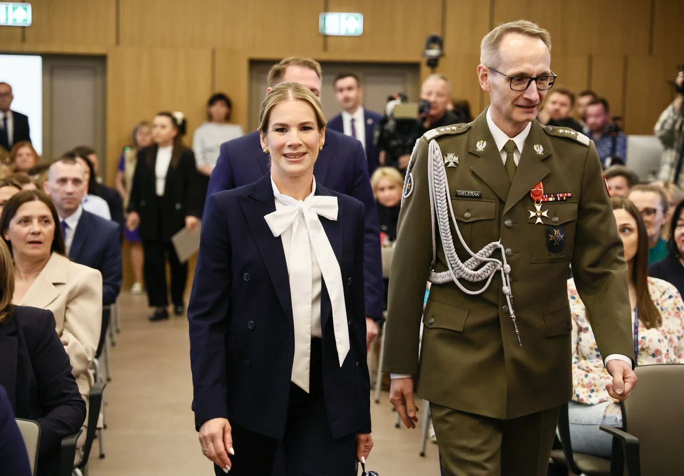 Kokardy pod szyją są hitem 2026 roku. Co jednak można było zrobić lepiej w tej stylizacji? Kobieta w eleganckim garniturze z białą kokardą na szyi i mężczyzna w mundurze wojskowym z odznaczeniami idą razem przez salę pełną siedzących ludzi.