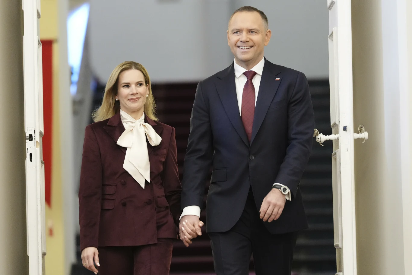 Pierwsza dama RP nie pierwszy raz sięgnęła po koszulę z wiązaniem w kokardę Elegancko ubrana para trzymająca się za ręce, zmierzająca razem przez jasny korytarz.