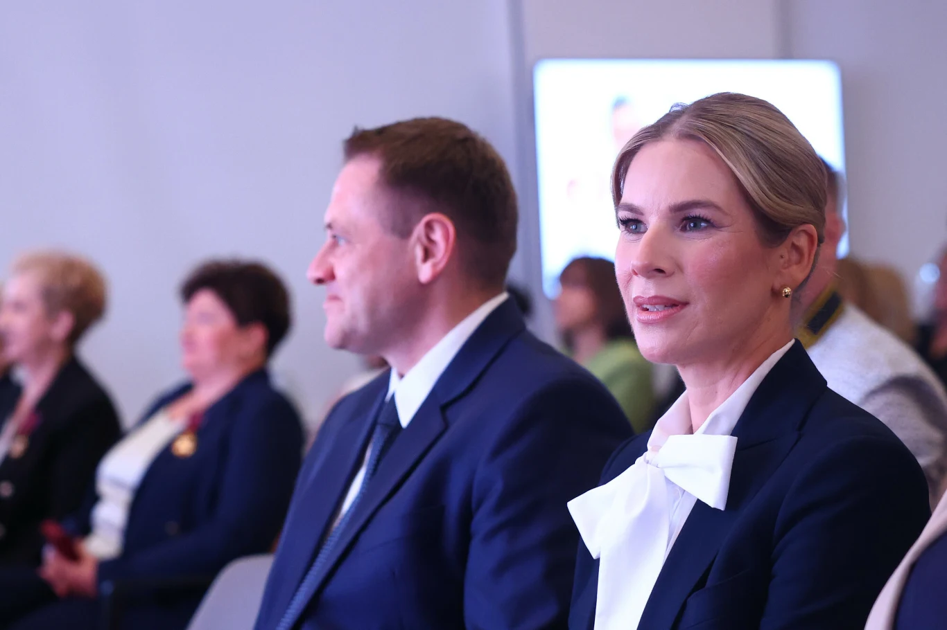 Biała kokarda mocno rzucała się w oczy na tle granatowego garnituru Kobieta w eleganckim granatowym garniturze z białą kokardą na szyi oraz mężczyzna w ciemnym garniturze siedzą obok siebie w pierwszym rzędzie podczas formalnego wydarzenia, w tle widoczne inne osoby ubrane w podobnym stylu.