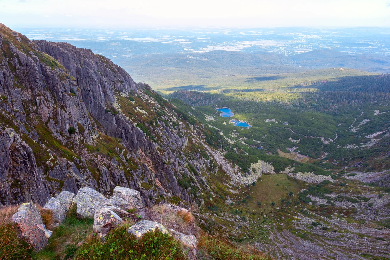 Widok na Śnieżne Kotły Stroma ściana skalna opadająca do szerokiej doliny z widocznym jeziorem pośrodku, otoczona zielenią lasów i łąk, w tle rozległa panorama gór i lasów sięgających po horyzont.