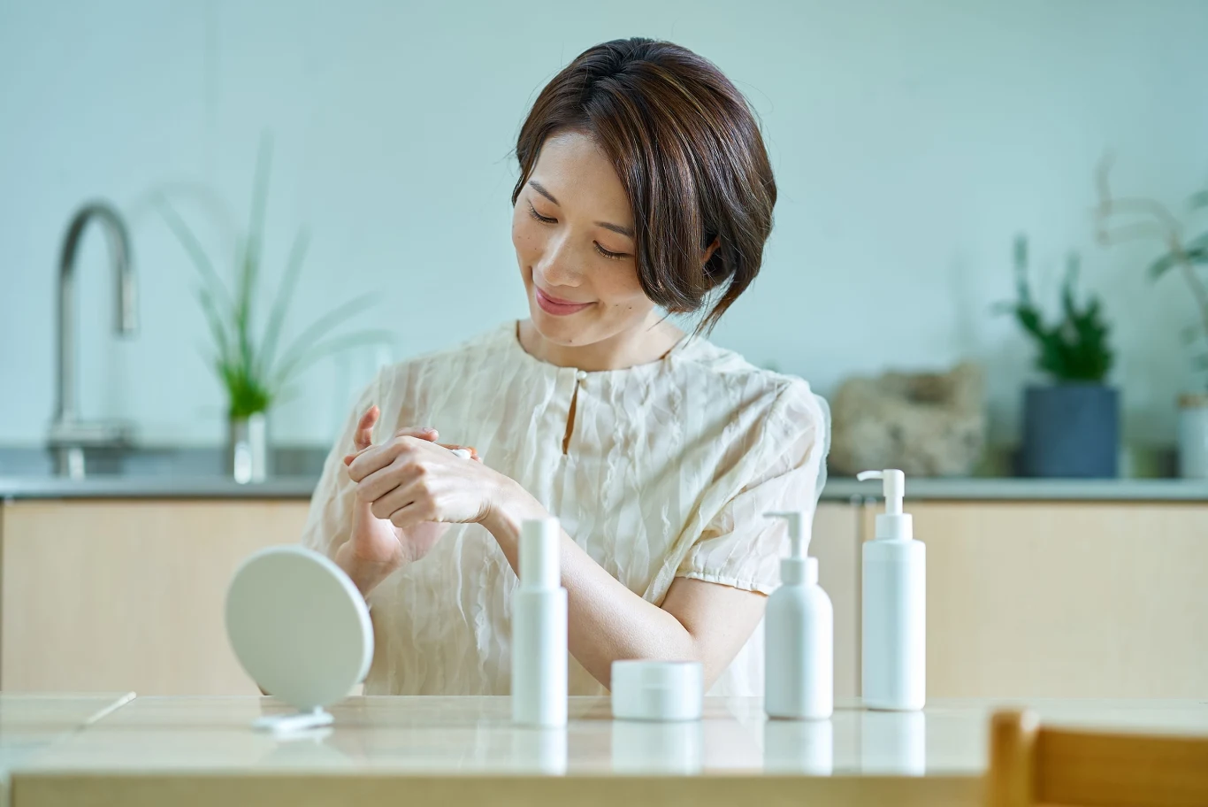 Kobieta nakładająca kosmetyk na dłonie, siedząca przy stole z lusterkiem i czterema białymi opakowaniami produktów pielęgnacyjnych.