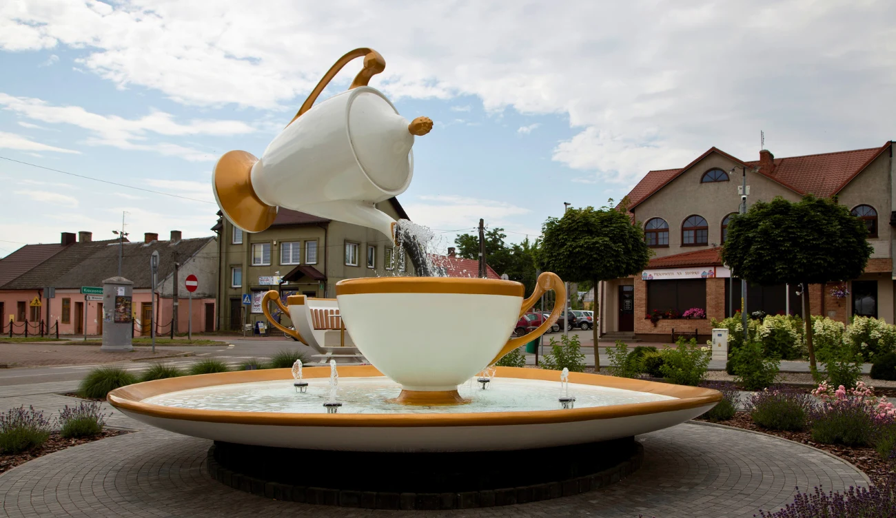 Jakie są atrakcje w Ćmielowie? Fontanna w kształcie filiżanki i dzbanka na Porcelanowym Rynku w Ćmielowie, otoczona budynkami i zielenią.