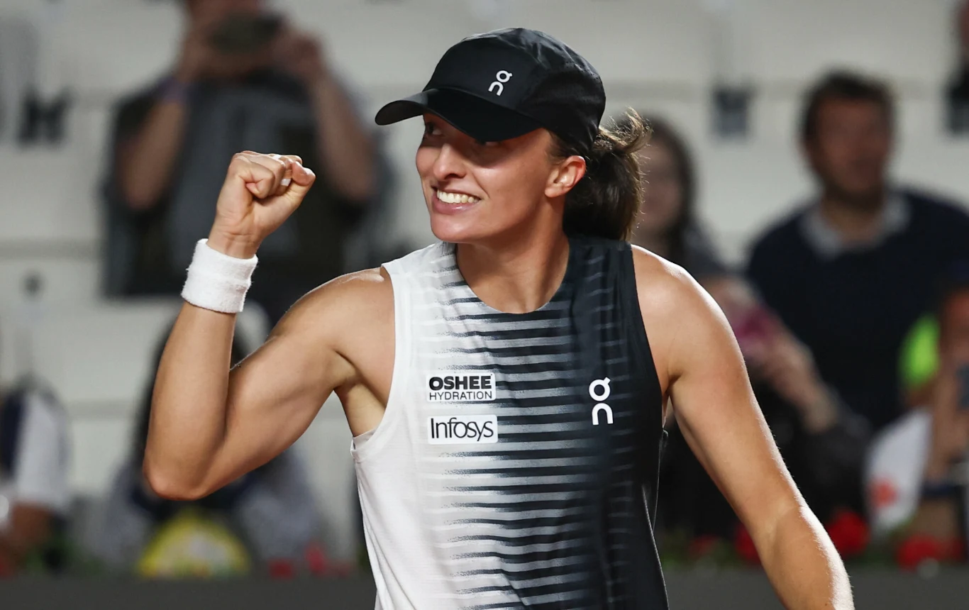 Uśmiechnięta tenisistka w sportowej koszulce i czapce z daszkiem unosi zaciśniętą pięść w geście triumfu na tle rozmytego tłumu.