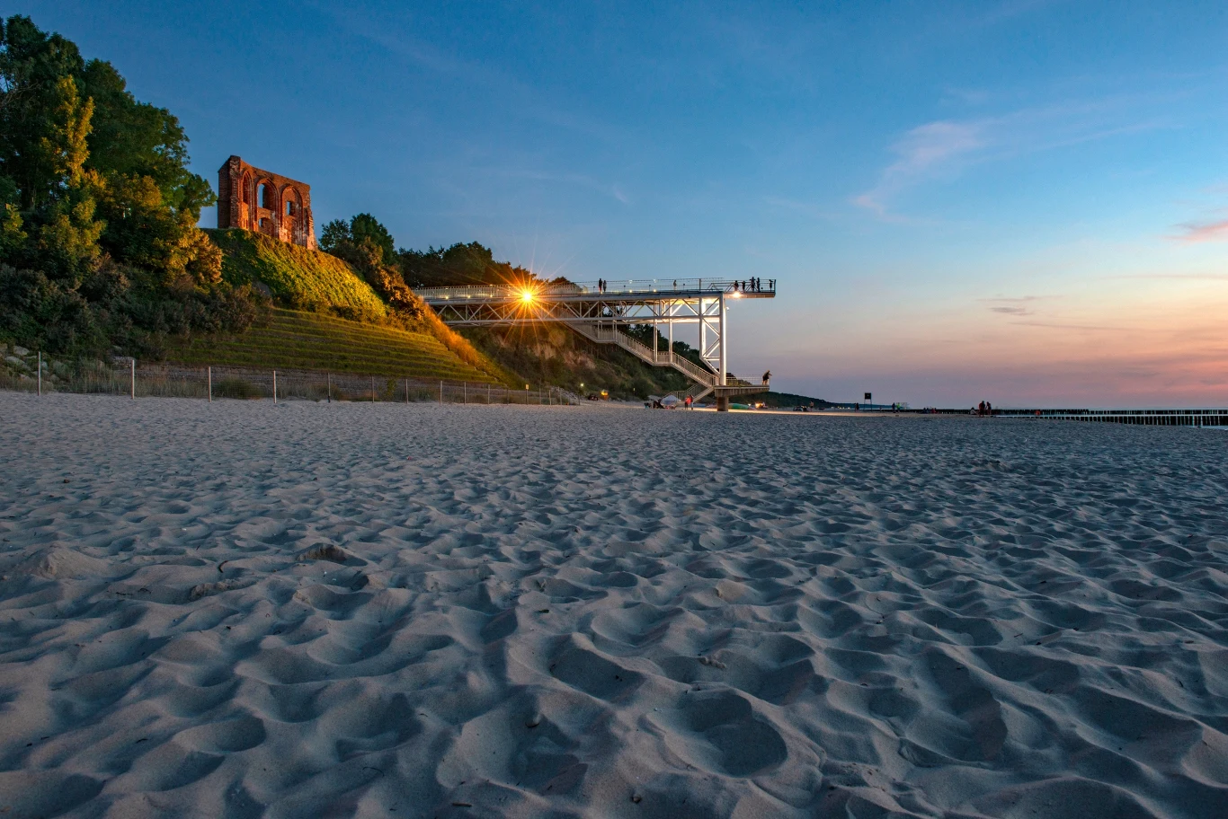 Platforma widokowa to jedno z najlepszych miejsc na podziwianie zachodów słońca Piaszczysta plaża z widokiem na klif, na którym znajduje się ruin gotyckiego kościoła oraz nowoczesna konstrukcja tarasu widokowego oświetlona światłem zachodzącego słońca.