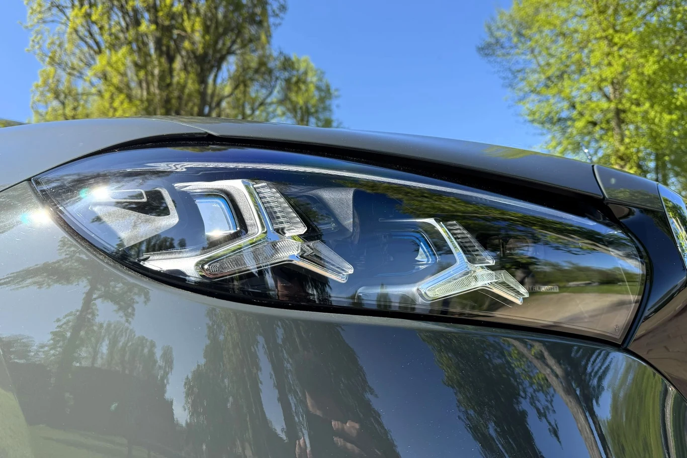 Reflektor nowoczesnego samochodu z wyrazistą linią diod LED, odbijający fragmenty otoczenia i nieba pośród zielonych drzew w słoneczny dzień.