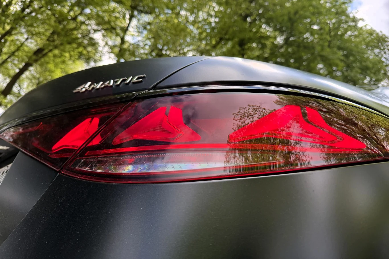Czerwone tylne światło samochodu marki Mercedes z oznaczeniem 4MATIC, odbijające zieleń otaczających drzew w połyskującym lakierze.