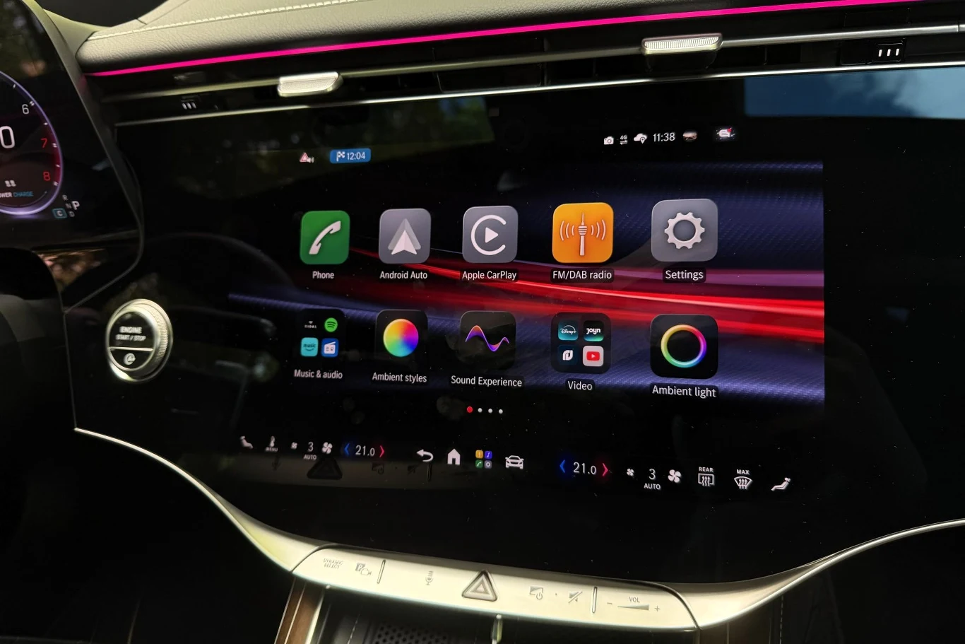 Nowoczesny ekran dotykowy systemu multimedialnego z kolorowymi ikonami aplikacji i funkcji, otoczony podświetleniem Ambient Light oraz elegancką czarną deską rozdzielczą z chromowanymi detalami.